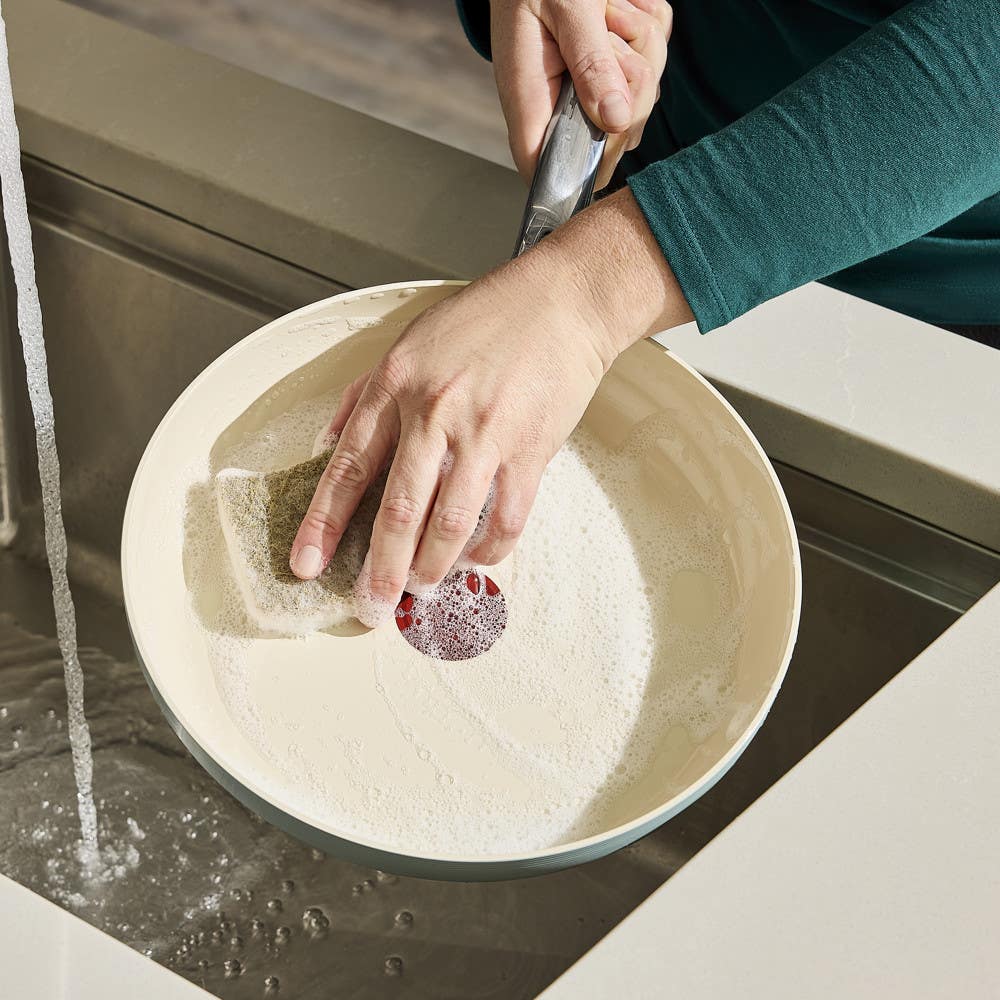 Hand washing a soapy ceramic frying pan under running water in a kitchen sink.