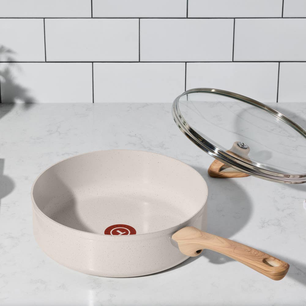 A beige frying pan with ceramic interior and wooden handle, with a lid on marble countertop