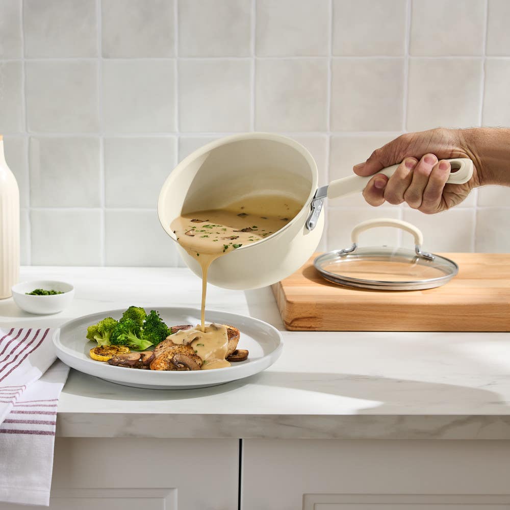 Hand pouring creamy sauce from beige nonstick saucepan over chicken, broccoli and mushrooms on plate