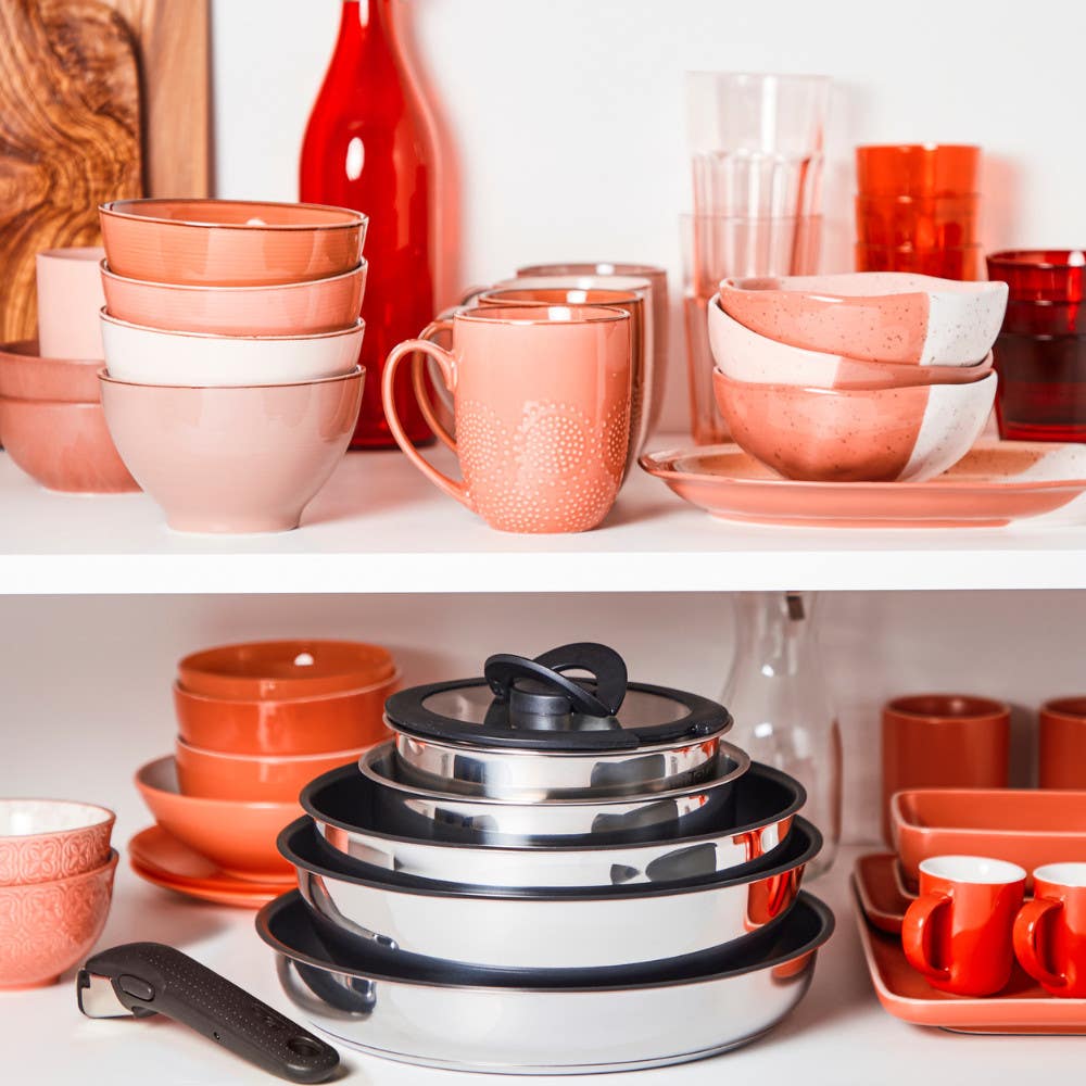 Stacked stainless steel cookware surrounded by orange and pink dishware in a cabinet