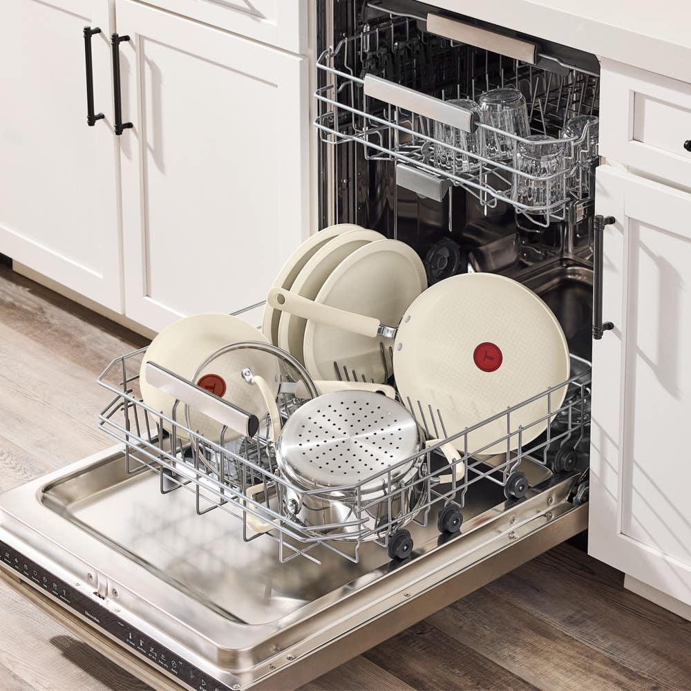 Open dishwasher loaded with beige nonstick cookware, glass lids and steamer insert after cleaning