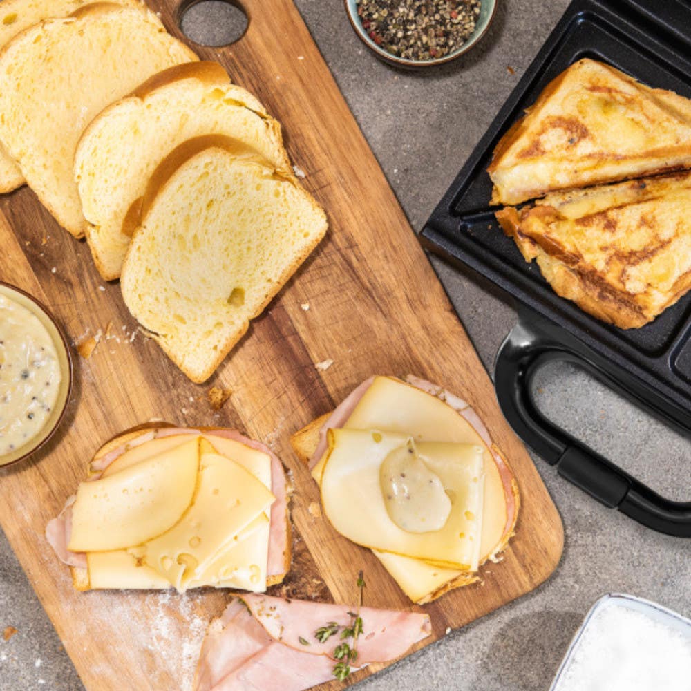 Sandwich maker nearby cooking a grilled sandwich, alongside sandwich ingredients with cheese and ham on a wooden board.