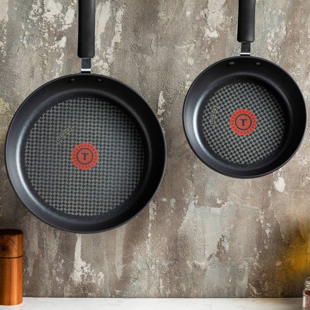 Two black nonstick frying pans with textured interiors and red heat indicators hang on a kitchen wall.