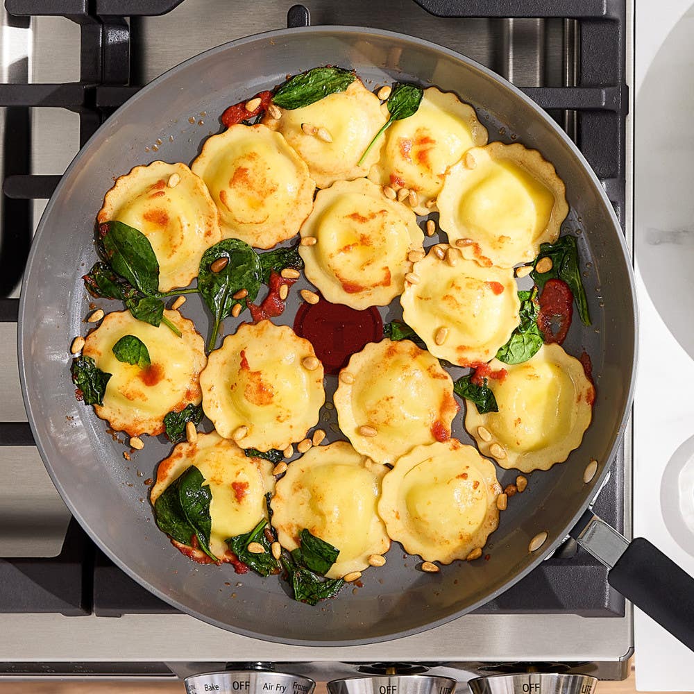 Ravioli with spinach and pine nuts in a ceramic skillet on a gas stovetop.