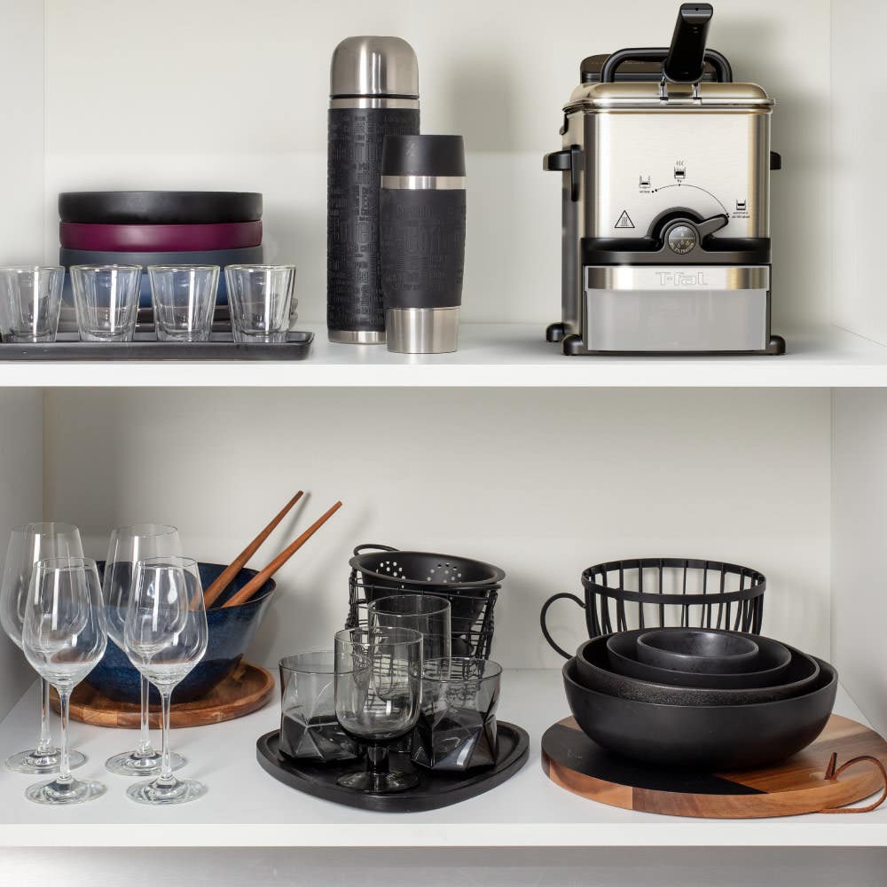 Shelf with household items: steel thermos, coffee machine, glasses, wine glasses, black ceramic bowls, and wooden trays. Various kitchen essentials neatly organized.