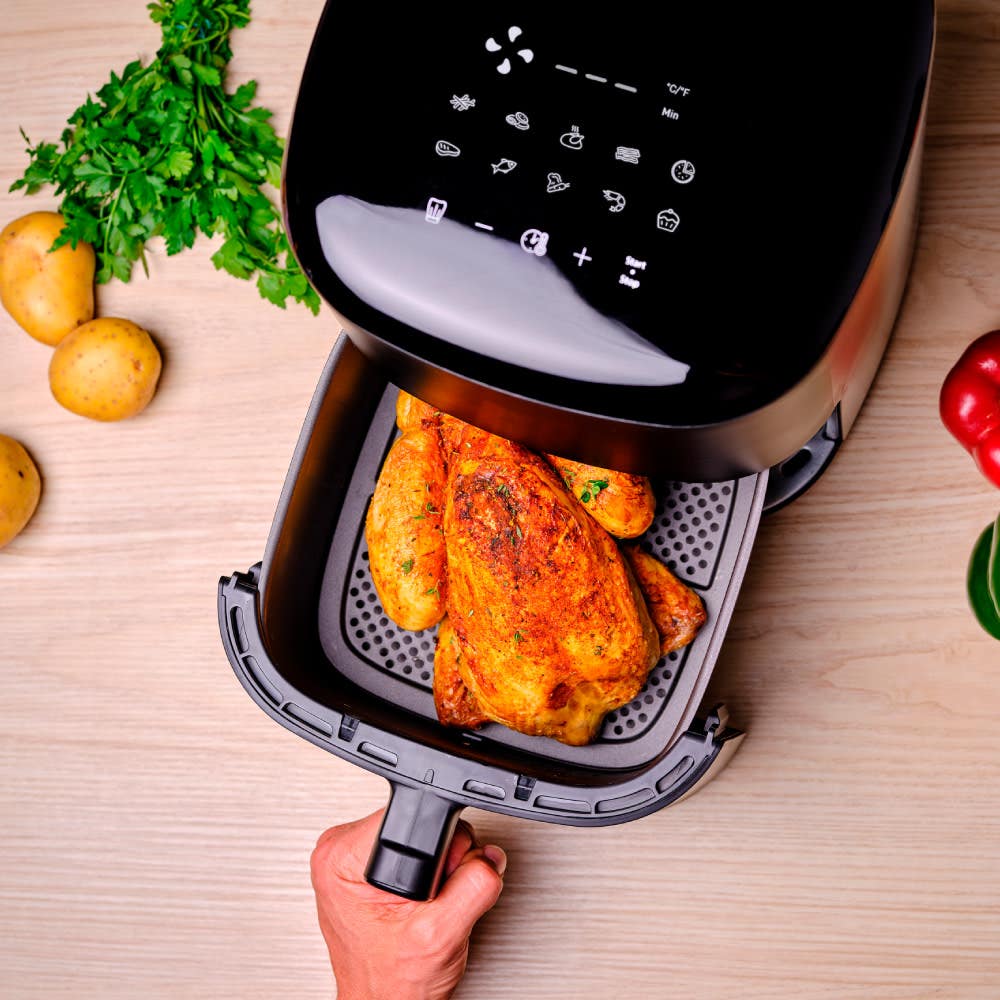 A hand holding the basket of a black air fryer with cooked chicken, surrounded by potatoes, parsley, and bell peppers on a wooden surface.