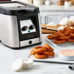 Deep fryer on a kitchen counter, surrounded by cooked onion rings and dipping sauce.