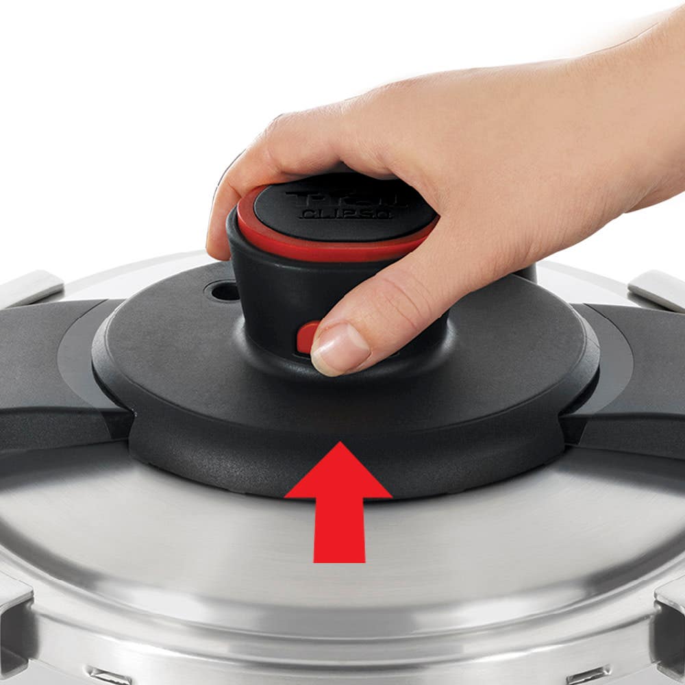 A hand pushes down on the central knob of a pressure cooker lid, with a red arrow pointing upward, indicating how to unlock the lid.