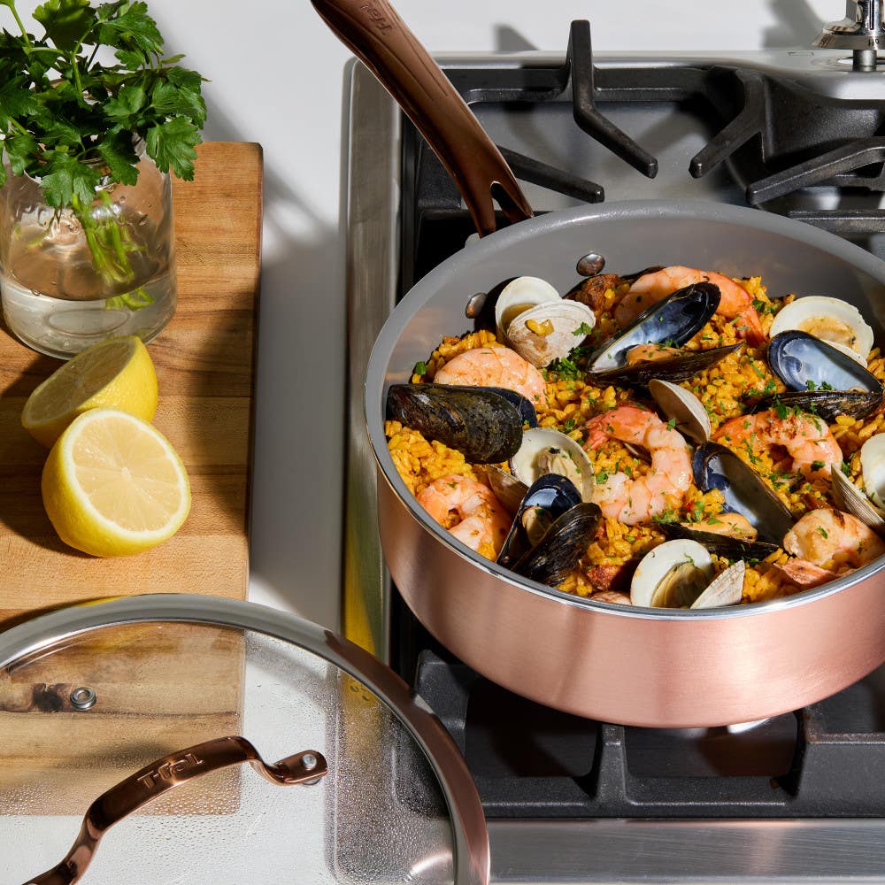 A jumbo cooker on a gas stove filled with seafood paella. Mussels, shrimp, and peas are visible. Sliced lemons sit on a wooden cutting board nearby.