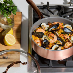 A jumbo cooker on a gas stove filled with seafood paella. Mussels, shrimp, and peas are visible. Sliced lemons sit on a wooden cutting board nearby.