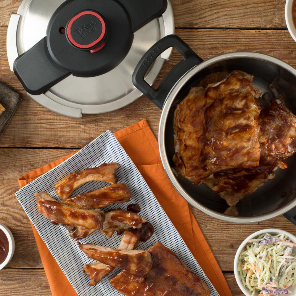 A pressure cooker filled with barbeque ribs is next to a plate of ribs and sides, set on a wooden table with an orange cloth.