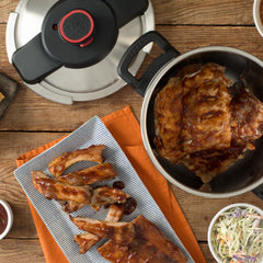 A pressure cooker filled with barbeque ribs is next to a plate of ribs and sides, set on a wooden table with an orange cloth.
