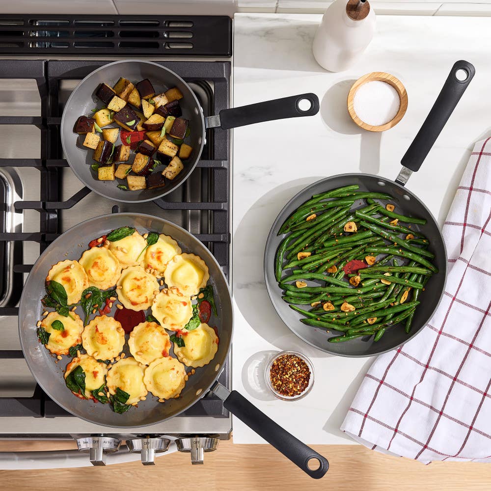 Overhead view of three ceramic pans cooking ravioli, eggplant, and green beans on a gas stove.
