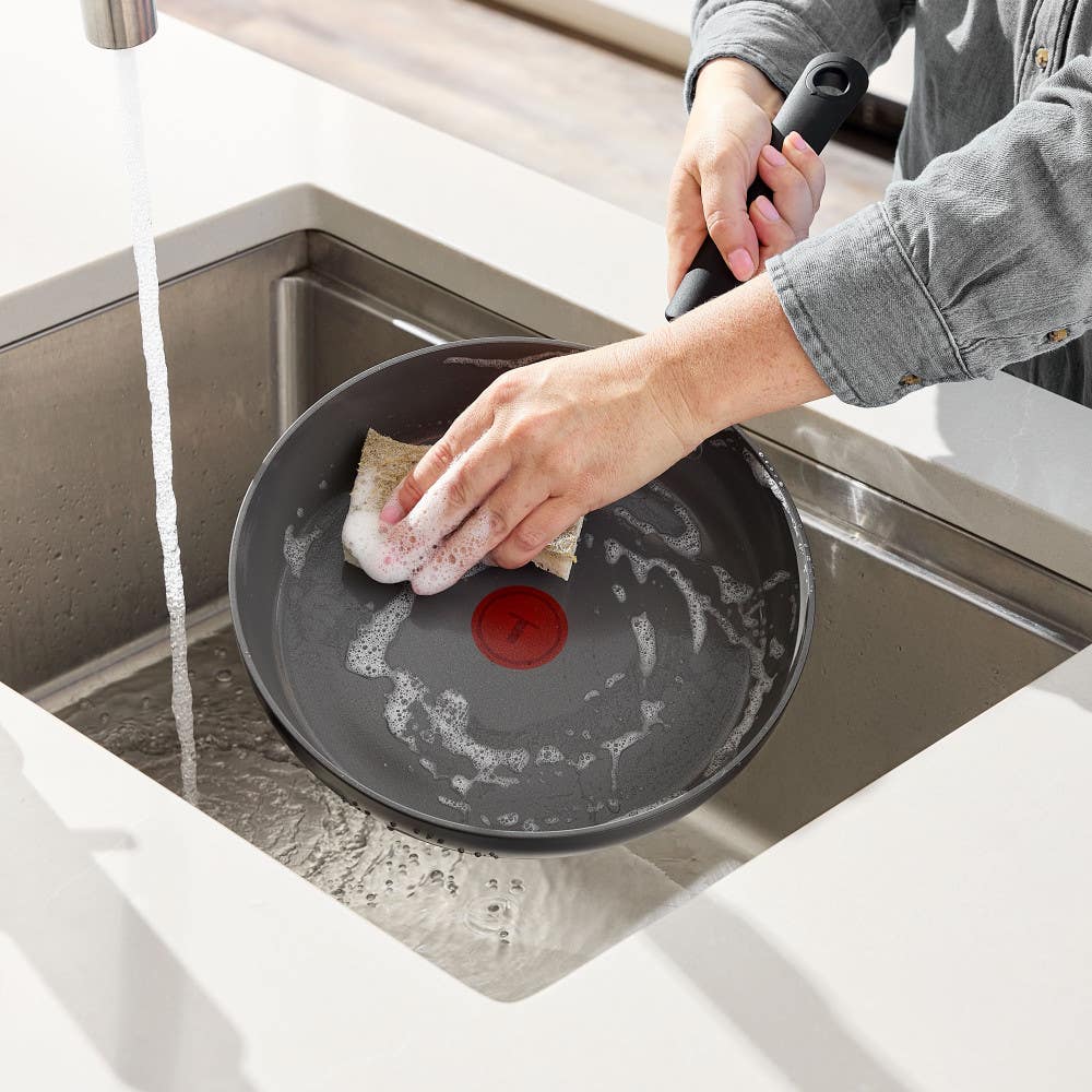 Person hand-washing a ceramic nonstick frying pan with a sponge in a kitchen sink.