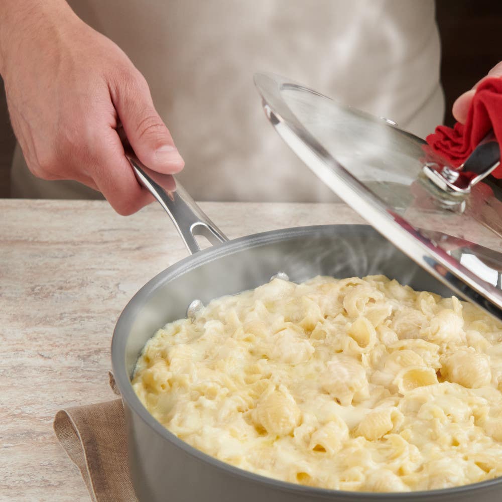 Hands lifting the lid off a steaming pan of pasta on a kitchen counter.