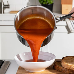 Pouring tomato soup from a stainless steel saucepan into a white bowl on a kitchen counter