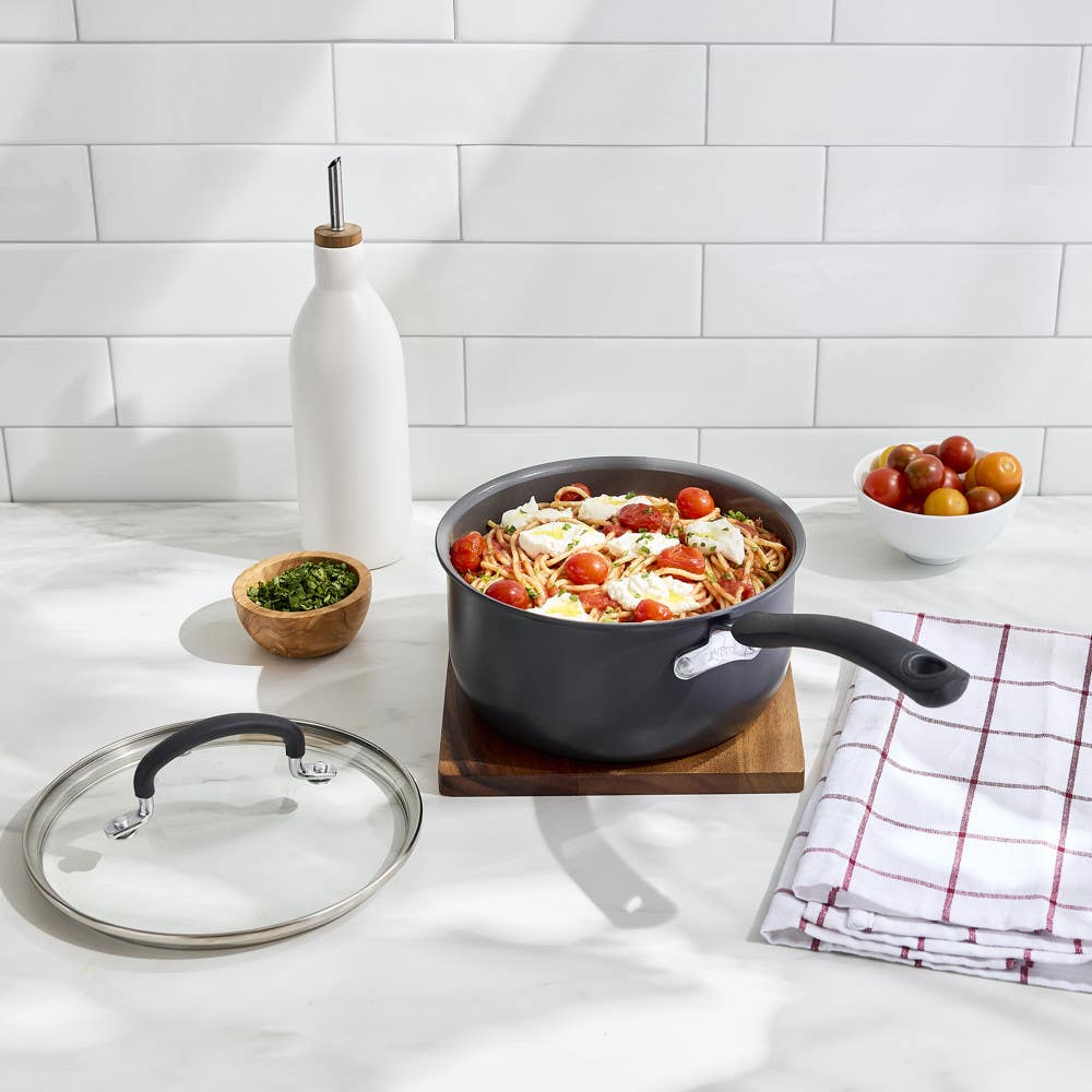 Ceramic saucepan with pasta, tomatoes, and cheese on a kitchen counter with ingredients nearby.