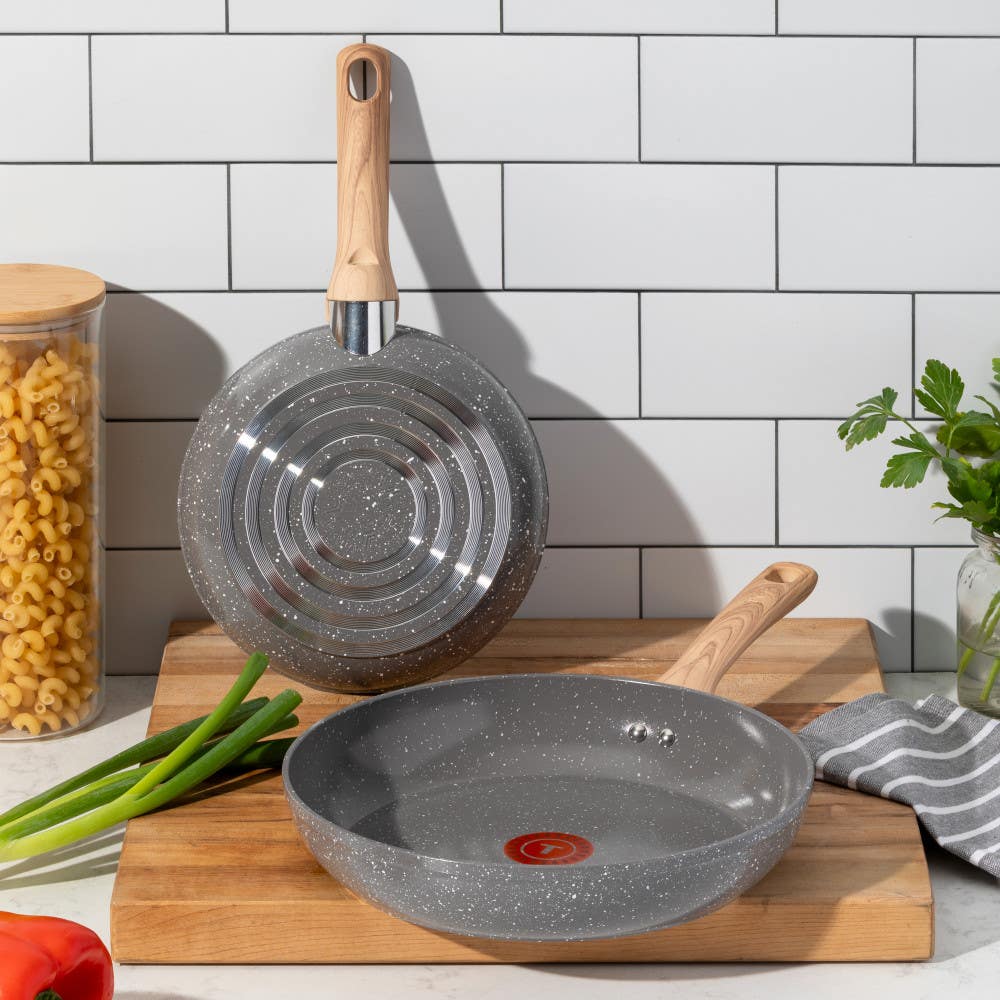 Two grey ceramic frypans with wooden handles on a kitchen counter with ingredients nearby