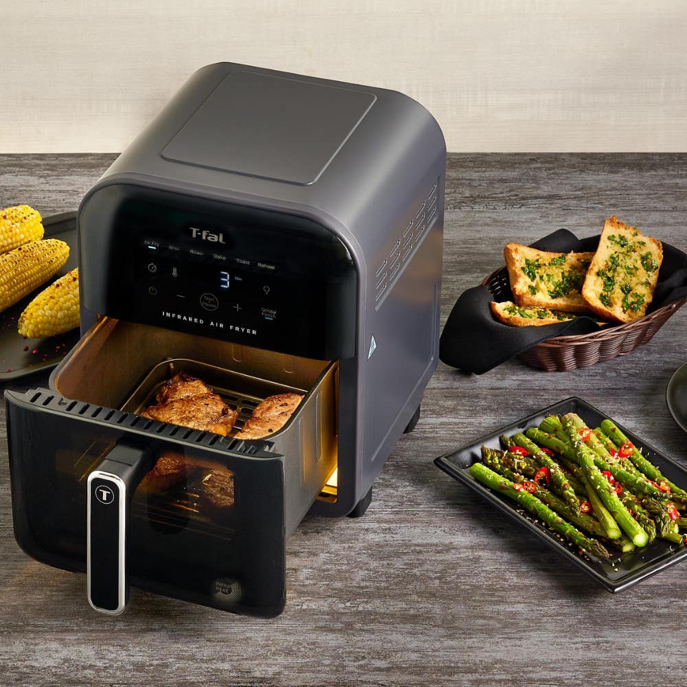 T-fal infrared air fryer on a kitchen counter with grilled corn, garlic bread, and asparagus. The air fryer basket contains well-cooked food