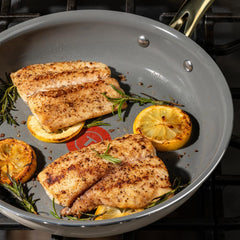 Two seasoned fish fillets cooking in a frying pan with sliced lemons and rosemary sprigs, placed on a gas stove.
