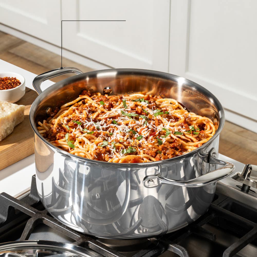 A stainless steel pot on a stove filled with spaghetti and sauce topped with herbs and cheese