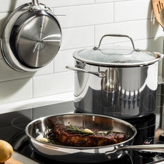 A stainless steel pot with a lid and a frying pan cooking steak on a stovetop in a kitchen