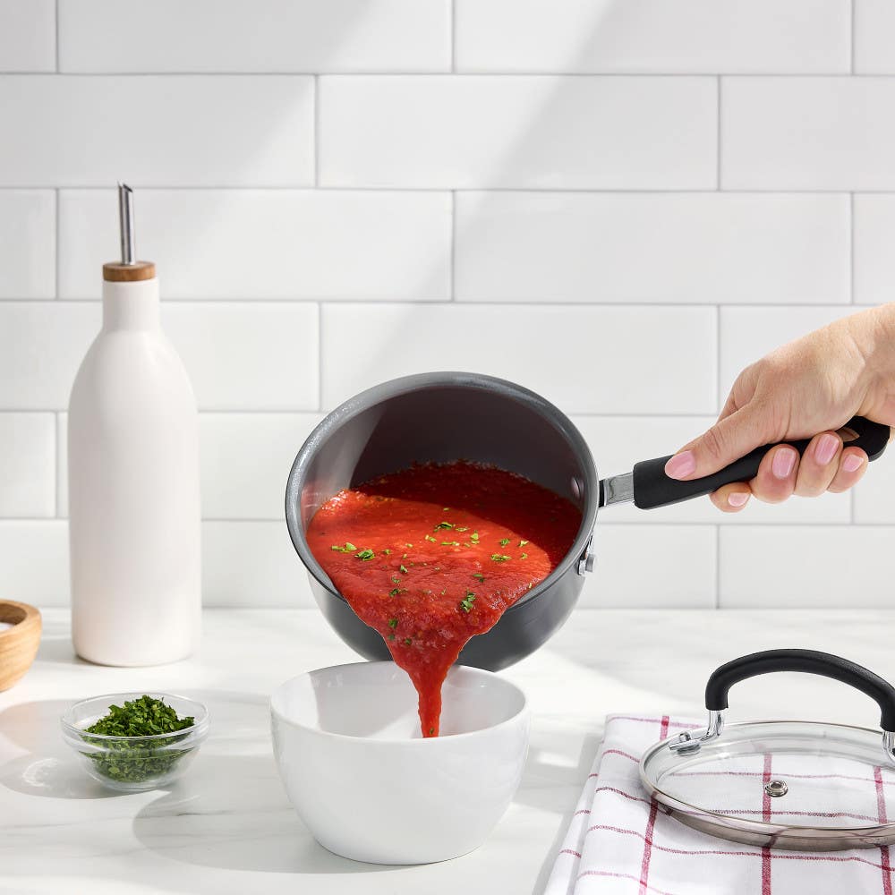 Hand pouring tomato soup from a ceramic saucepan into a white bowl on a kitchen counter.
