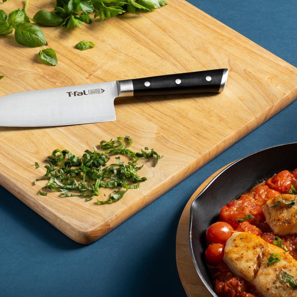 T-fal santoku knife on a wooden cutting board with chopped herbs next to a pan of chicken.