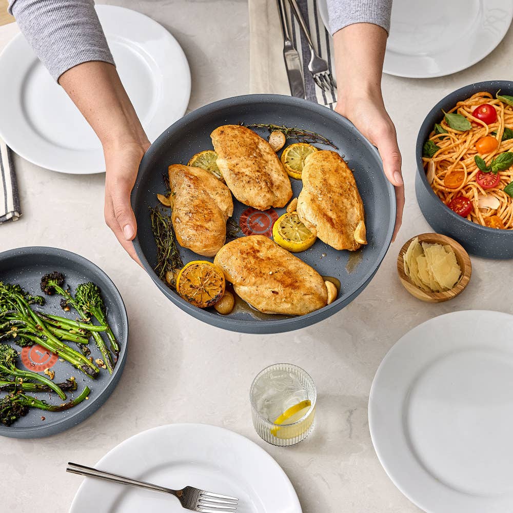 Hands hold a gray pan of roasted chicken and lemons, surrounded by dishes and empty plates