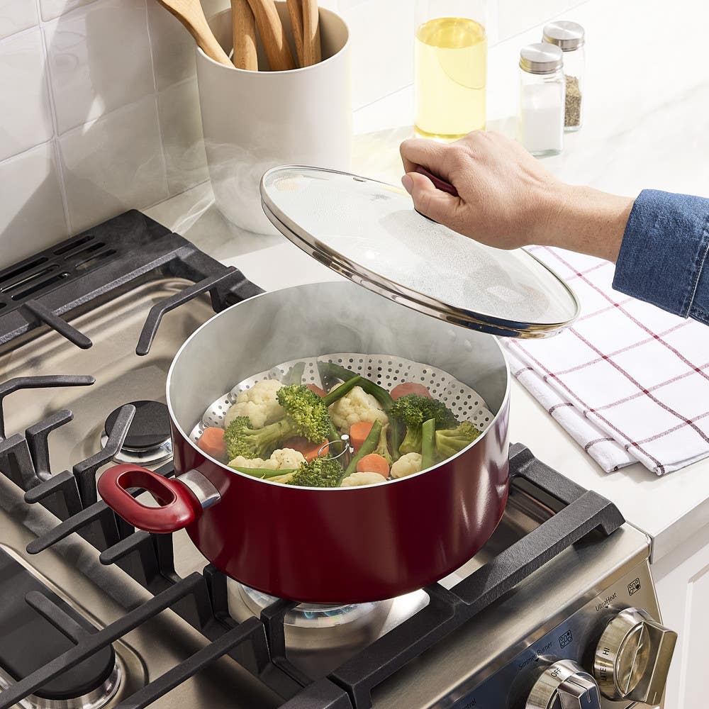 Hand lifting glass lid from red steamer pot with mixed vegetables on gas stove