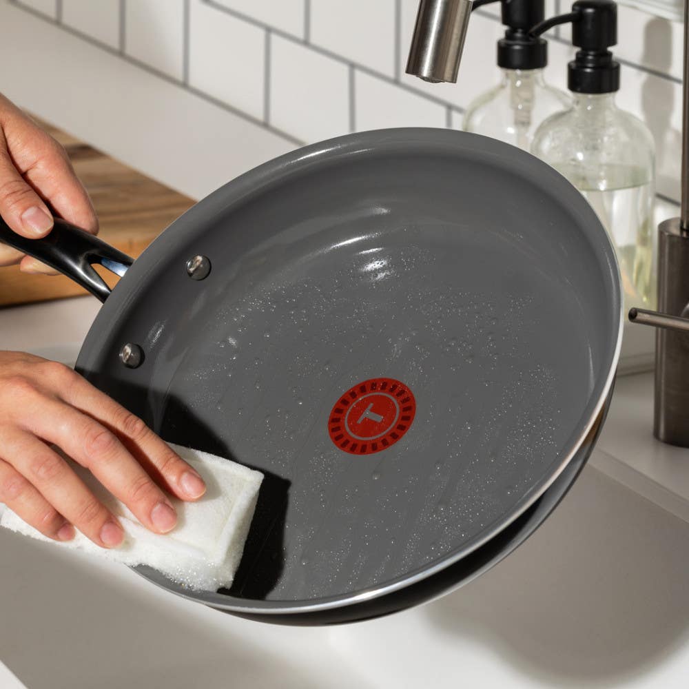 Person cleaning the inside of a pan with a white sponge, showing T-fal's red heat indicator in the center on a kitchen counter.