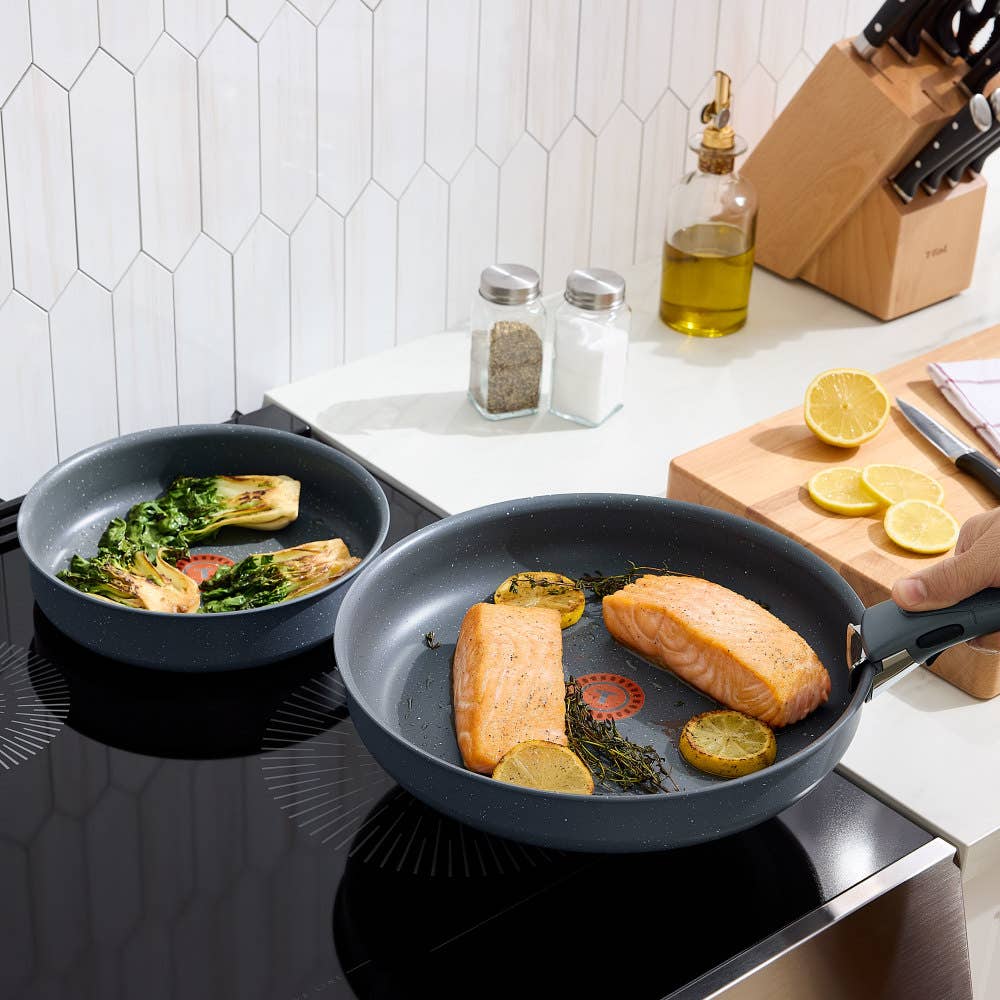 Two gray frying pans with detachable handles on an induction stove, cooking colorful meals