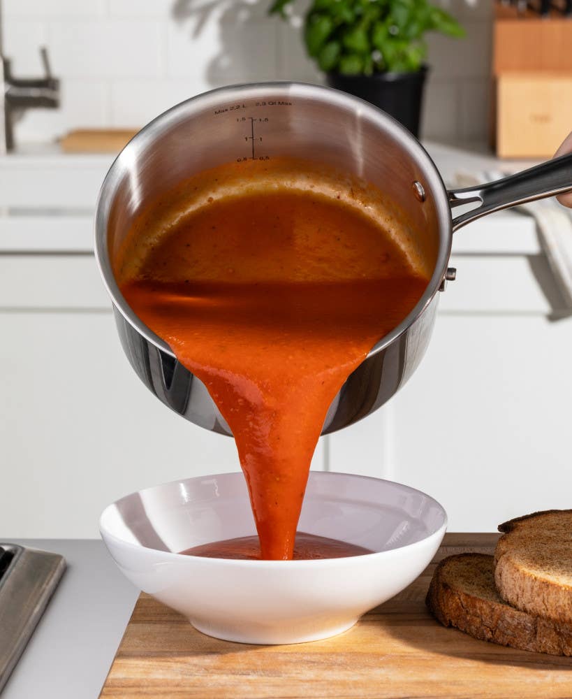 Pouring tomato soup from a stainless steel saucepan into a white bowl on a kitchen counter