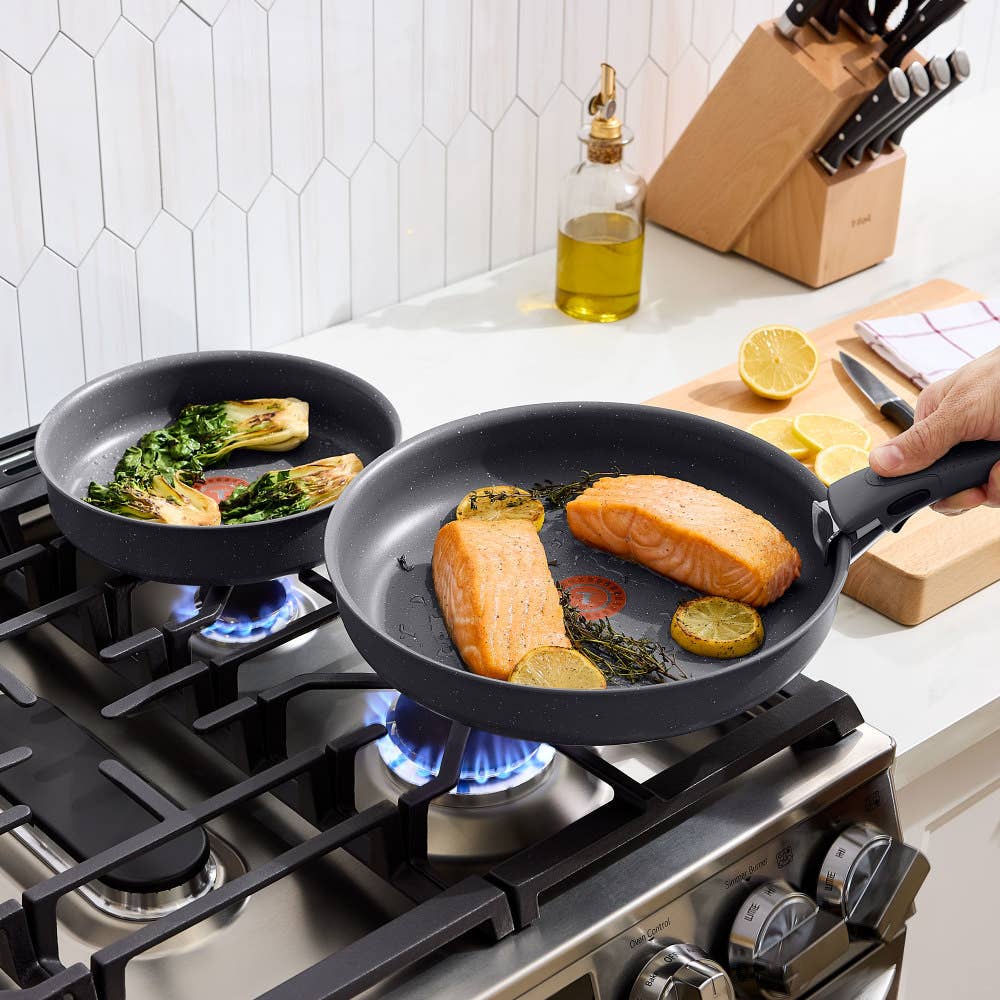 Two gray frying pans with detachable handles on a gas stove, cooking fish and vegetables