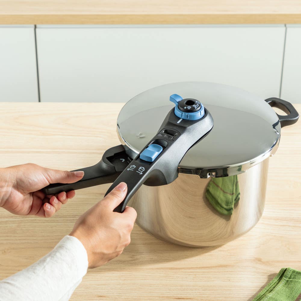 Hands closing a stainless pressure cooker lid with black and blue handles on wood counter