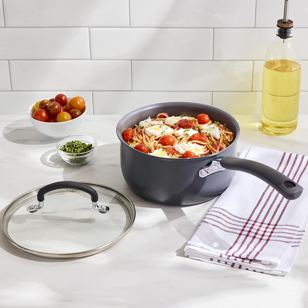 Nonstick saucepan filled with pasta and tomatoes, lid and towel beside, on a kitchen counter.