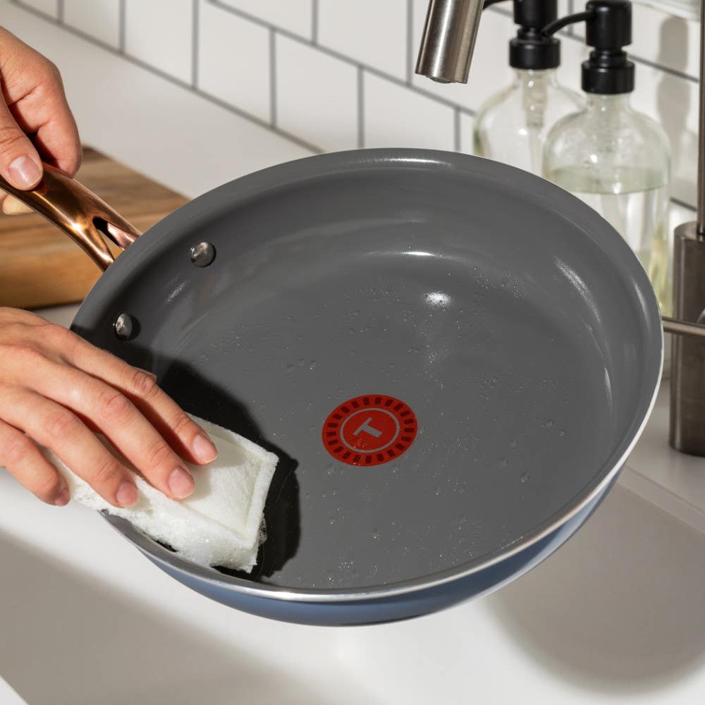 Person cleaning the inside of a pan with a white sponge, showing T-fal's red heat indicator in the center on a kitchen counter.