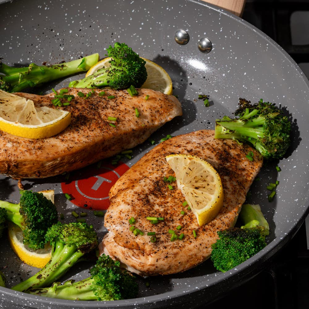 Two seasoned chicken breasts with lemon slices and broccoli cooking in a ceramic frypan