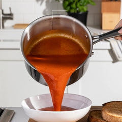 Pouring tomato soup from a stainless steel saucepan into a white bowl on a kitchen counter