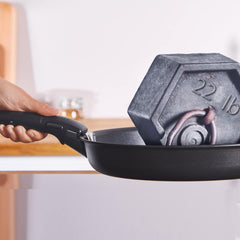A black frying pan holding a grey 22 lb weight, with a hand grasping the handle