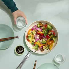 Ceramic frypan filled with colorful stir-fried vegetables on a table with plates, chopsticks, and glasses.