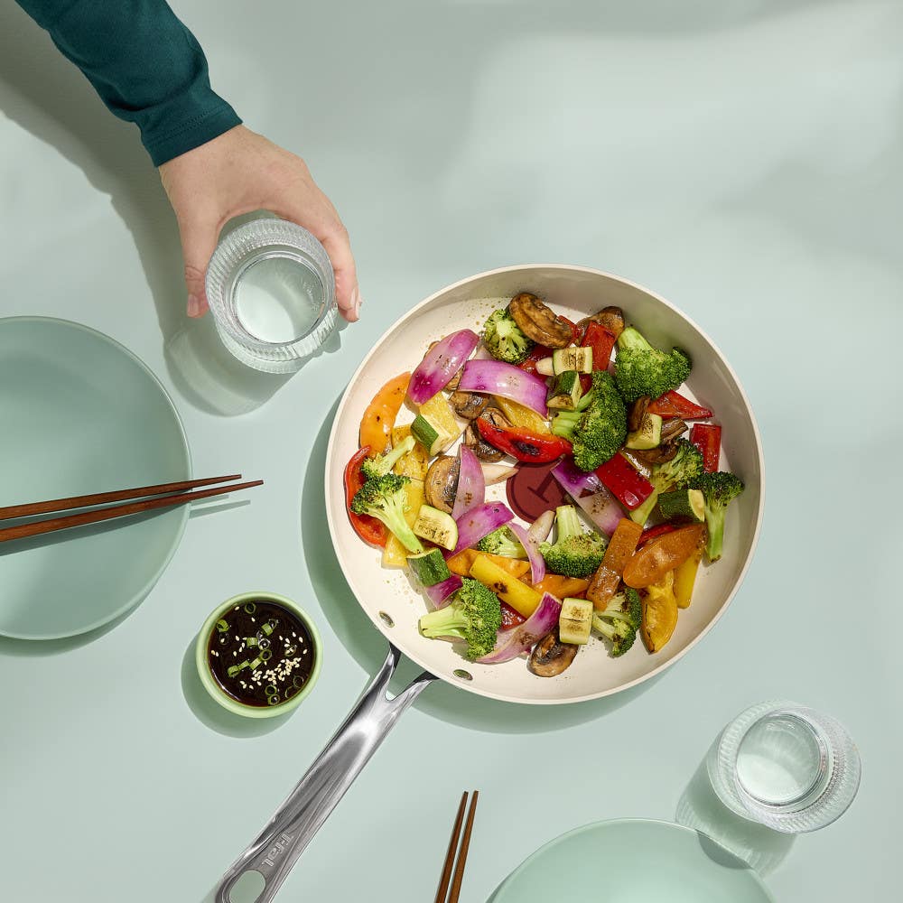 Ceramic frypan filled with colorful stir-fried vegetables on a table with plates, chopsticks, and glasses.