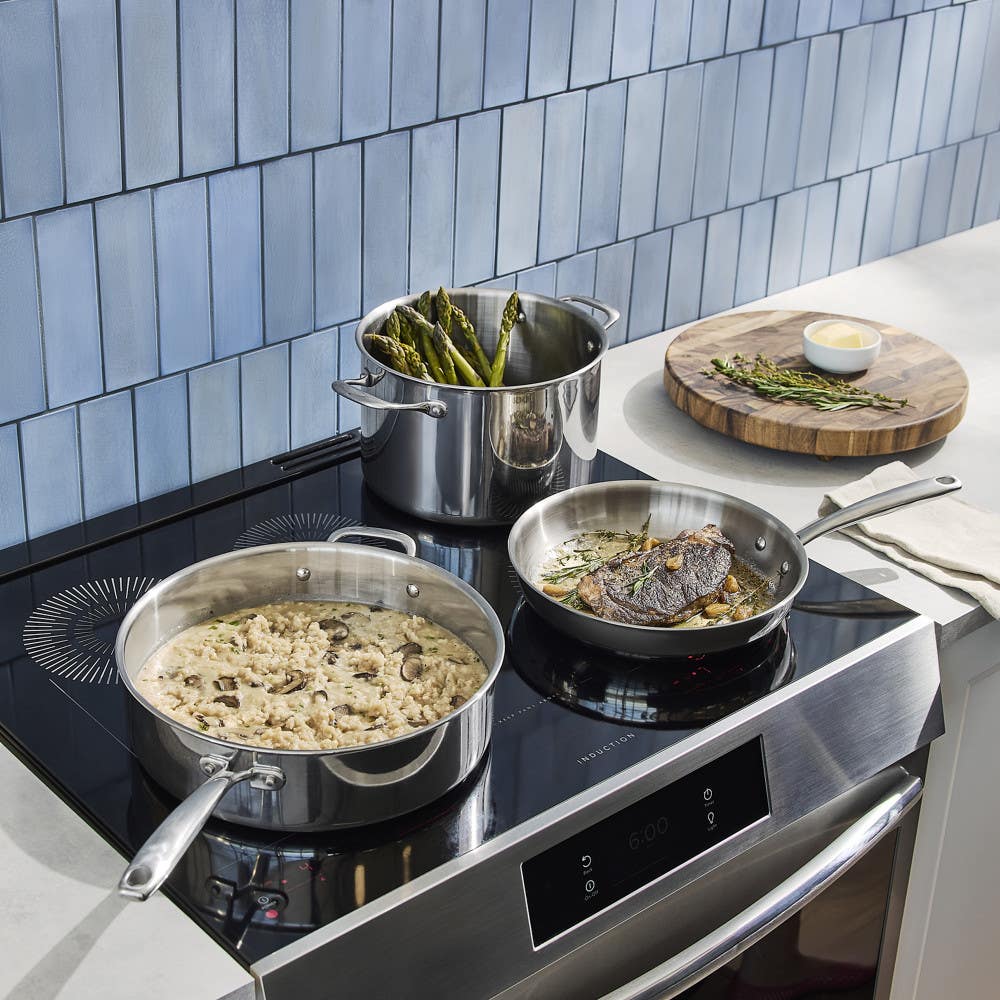 Image of three stainless steel pots and pans on an induction stove top. One pot holds asparagus, one pan has creamy risotto with mushrooms, and another pan cooks a piece of fish with herbs. There is a wooden cutting board with herbs and a small bowl of bu