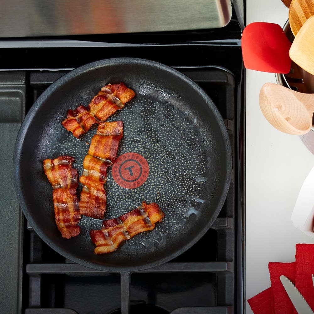 Black non-stick frying pan with T-fal logo cooking four strips of bacon on a stove