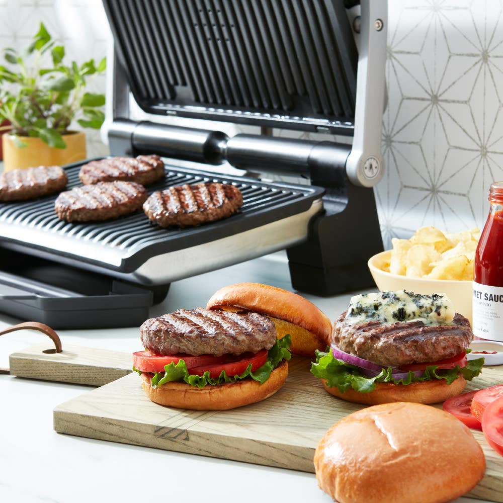 OptiGrill cooking burger patties with prepared burgers, buns, and condiments on a kitchen counter.