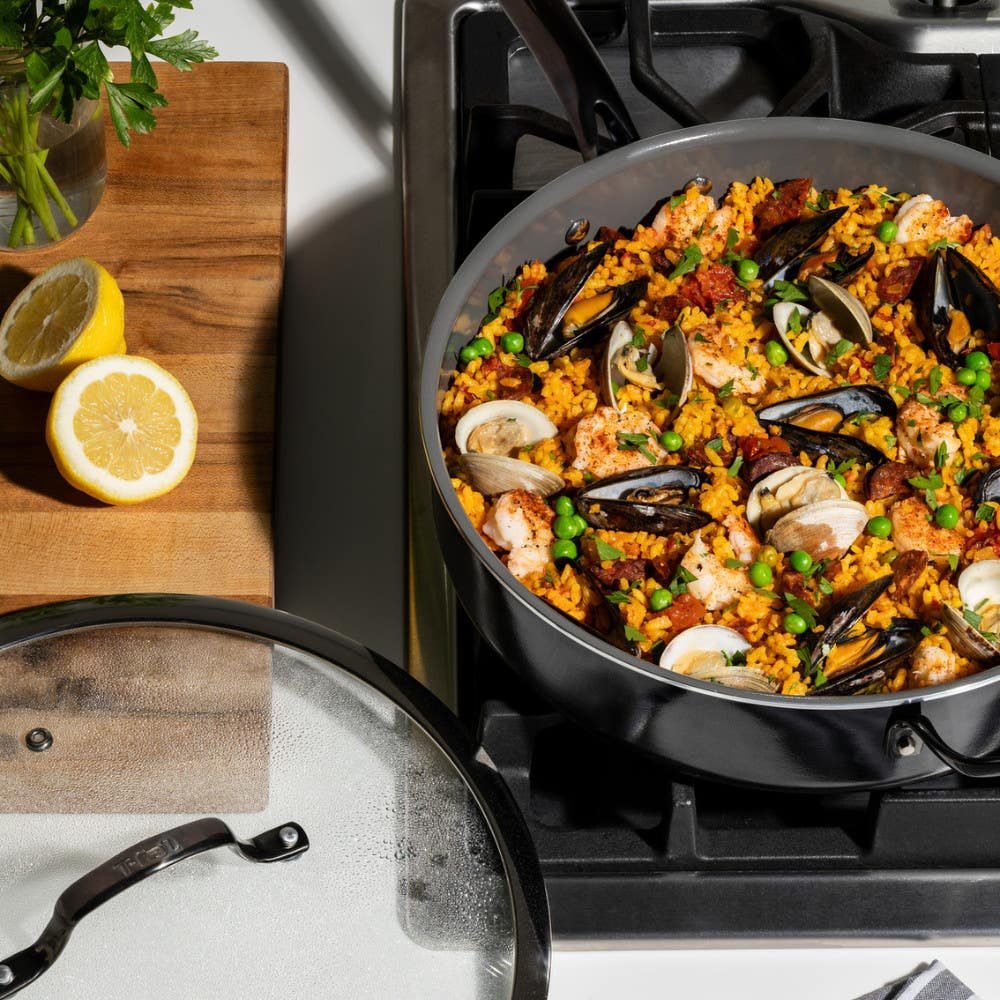 A jumbo cooker on a gas stove filled with seafood paella. Mussels, shrimp, and peas are visible. Sliced lemons sit on a wooden cutting board nearby.