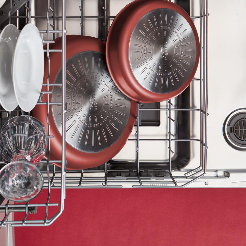 Two red frying pans placed upside down in a dishwasher along with glass cups and plates
