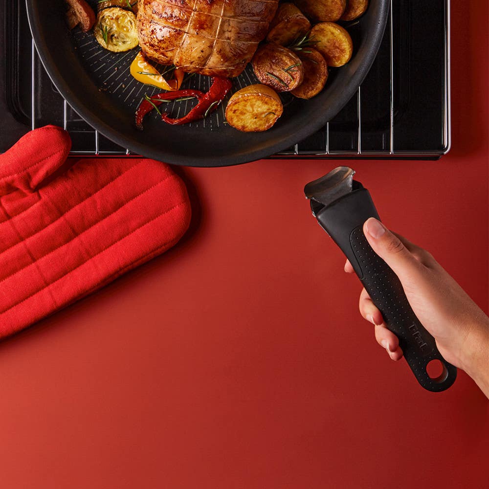 Black pan with roasted food, a red oven mitt, and a hand holding a blue detachable handle