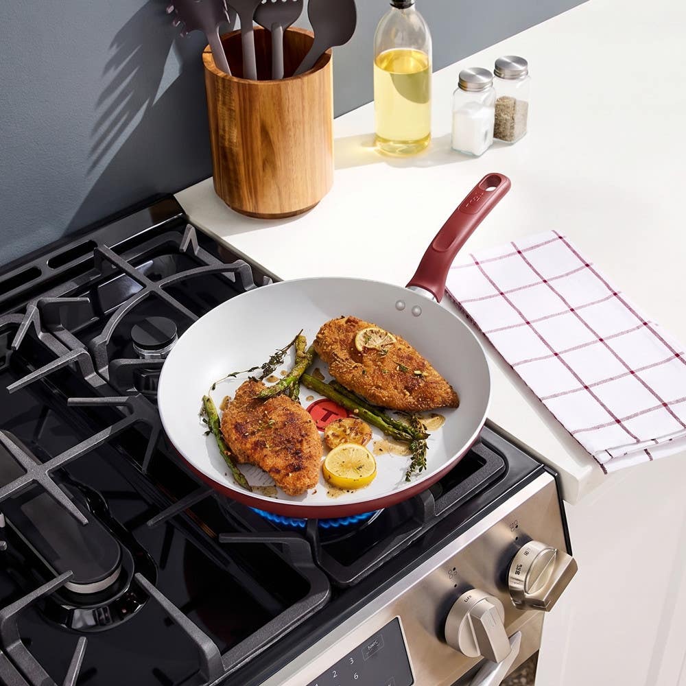 Red frypan cooking two seasoned chicken breasts with vegetables on gas stove