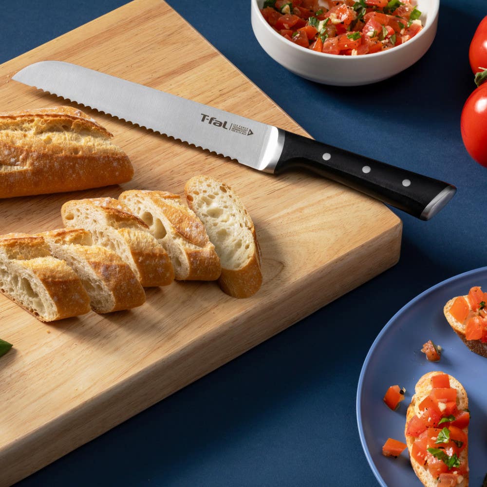 Sliced baguette with serrated knife, tomatoes and toppings nearby.